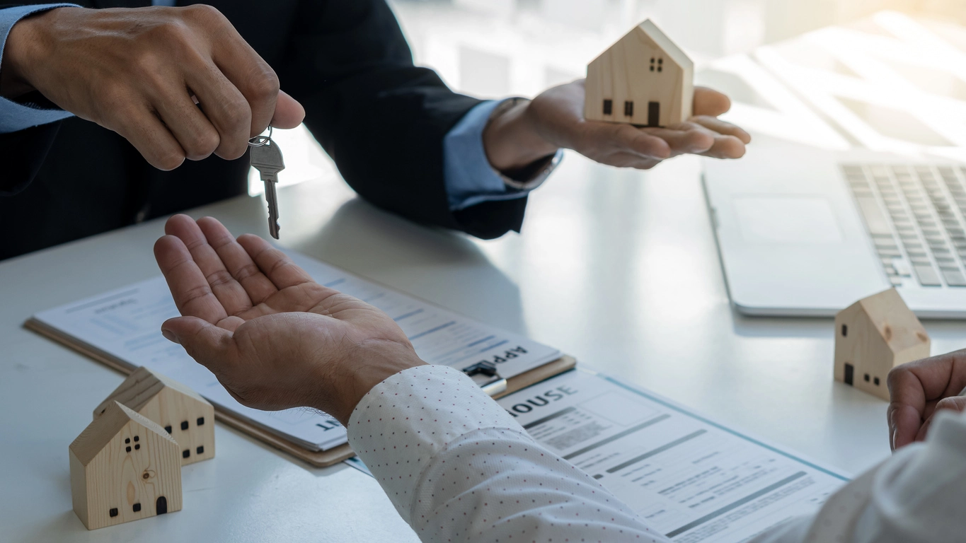 Un agent immobilier remet des clés à une personne, symbolisant la délégation de la gestion locative Angers. Des miniatures de maisons et des documents administratifs sont visibles sur un bureau