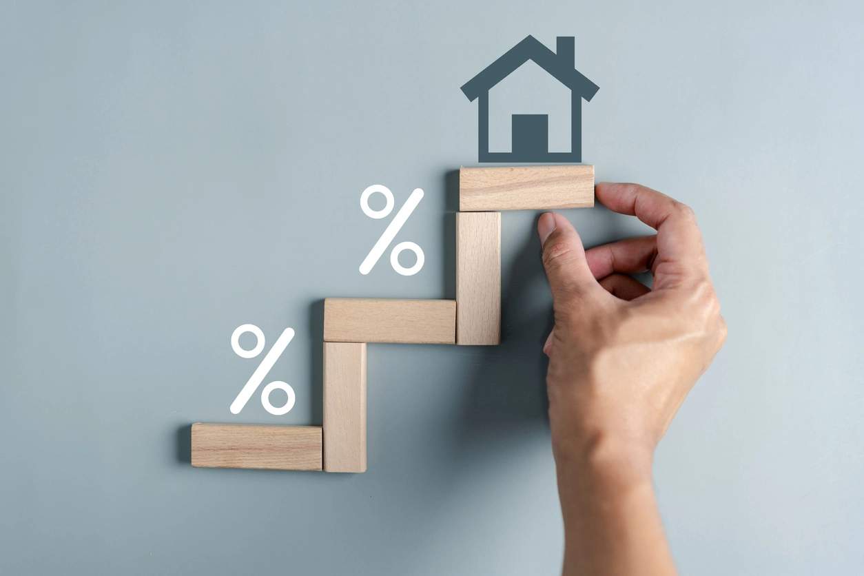 Une main place un bloc en bois sur une structure en escalier symbolisant l’investissement immobilier. Des pourcentages et une icône de maison illustrent la croissance.