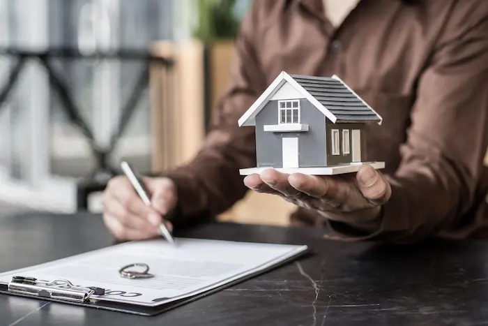Personne en chemise brune tenant une maquette de maison et un stylo devant contrat, image illustrant le rôle du courtier crédit immobilier 