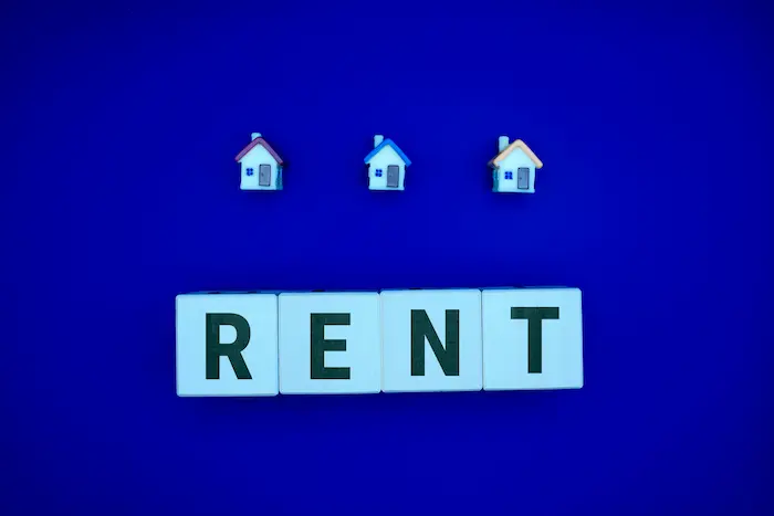 Trois petites maisons alignées au-dessus de cubes blancs formant le mot RENT, image illustrant l’encadrement des loyers à Lyon.