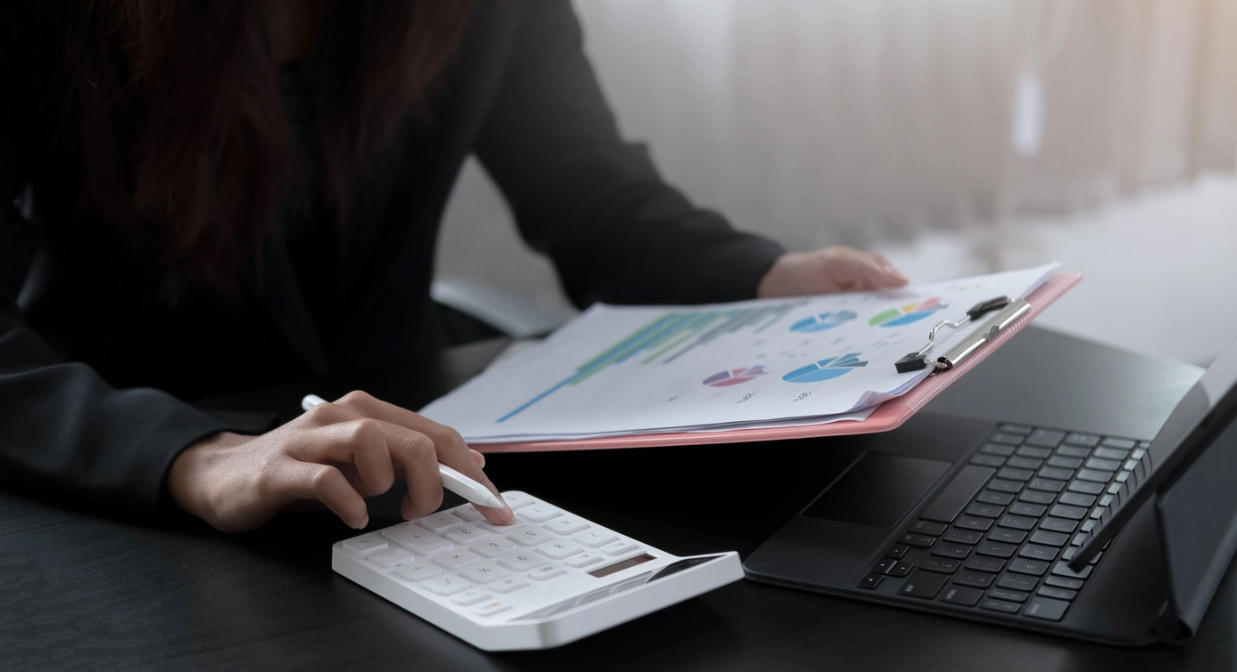 Sur cette image, une personne est concentrée sur le calcul des frais de gestion locative. Elle tient une calculatrice et examine attentivement des documents ornés de graphiques en cercles