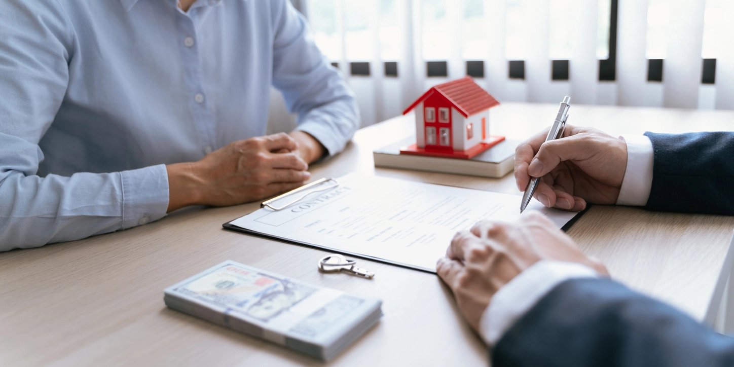 Deux personnes en discussion pour la signature d'un contrat immobilier, avec une clé, une liasse de billets et une maquette de maison sur un bureau en bois.