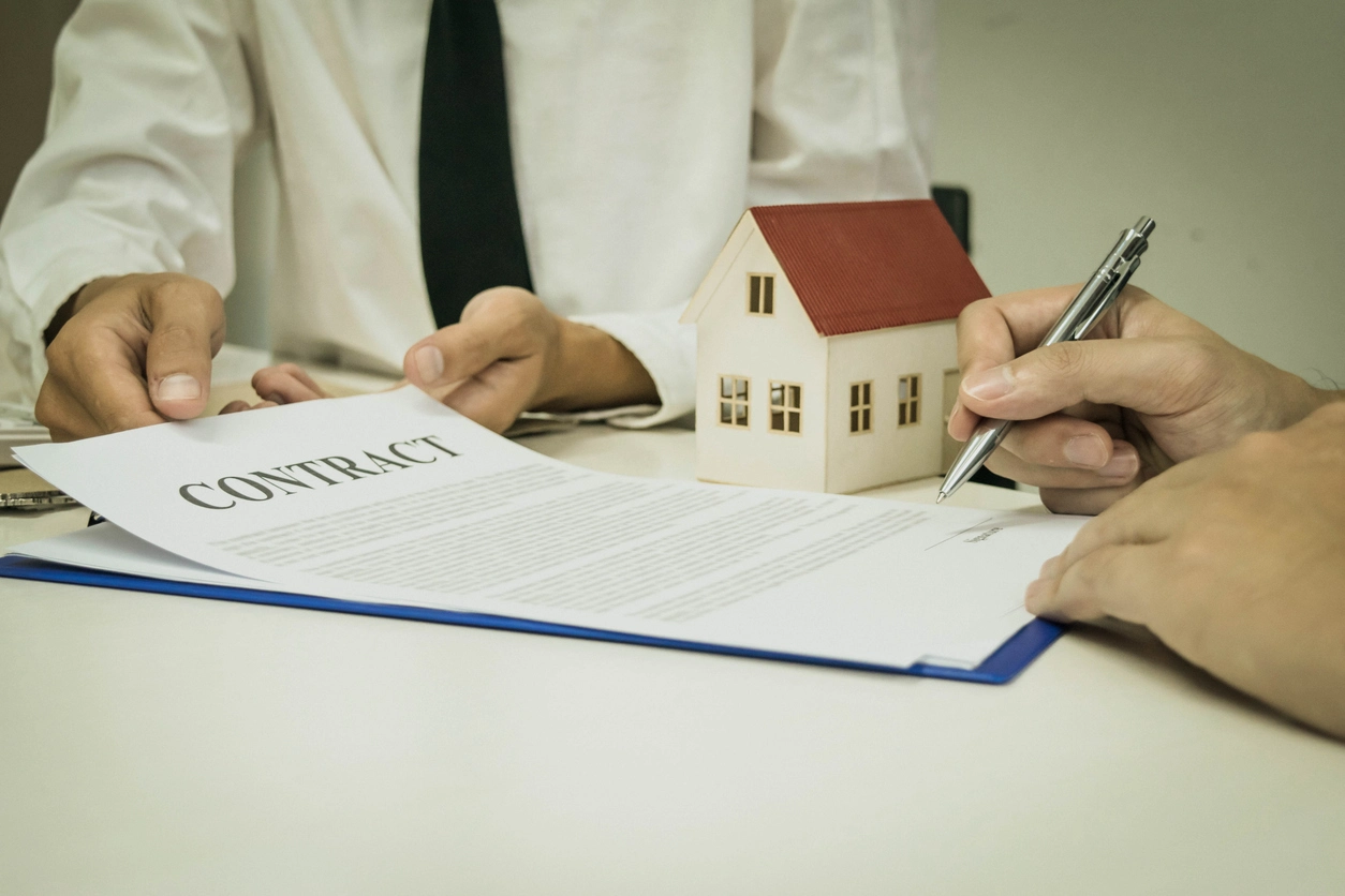 Signature d'un contrat de gestion locative entre un propriétaire et un agent immobilier, avec une maquette de maison en arrière-plan symbolisant l'investissement immobilier