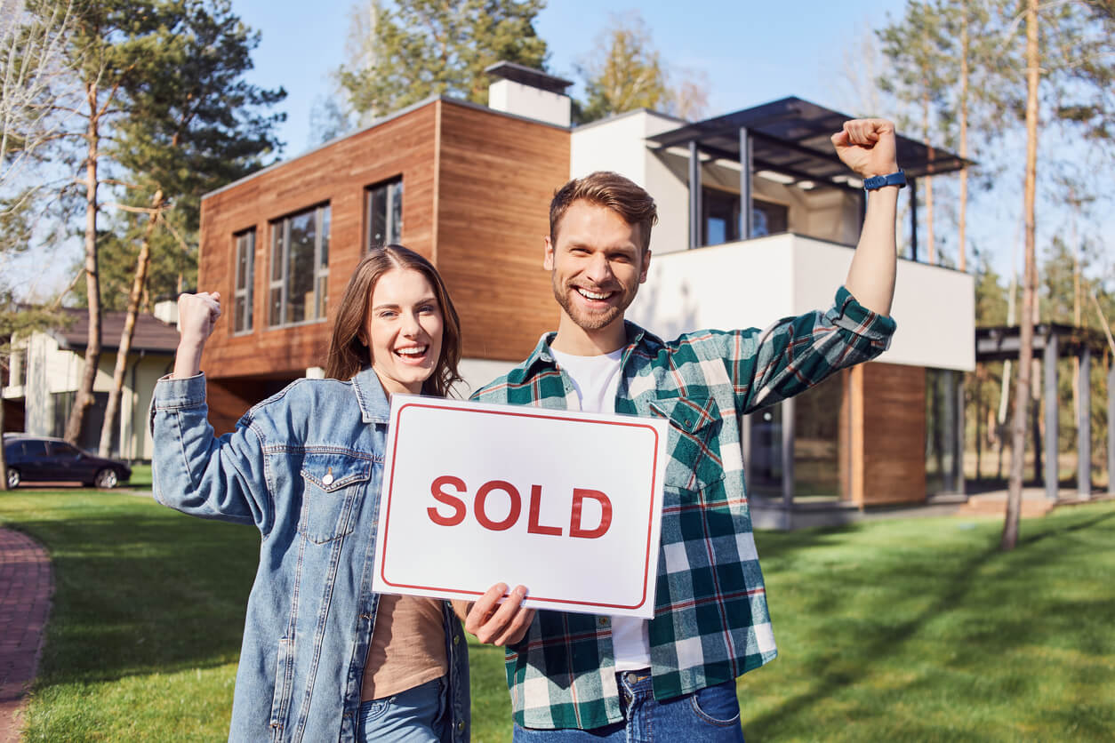 Un couple heureux pose devant leur nouvelle maison moderne, avec un panneau "Vendu" en évidence. La maison, floue en arrière-plan, symbolise leur succès dans un investissement locatif Tourcoing.