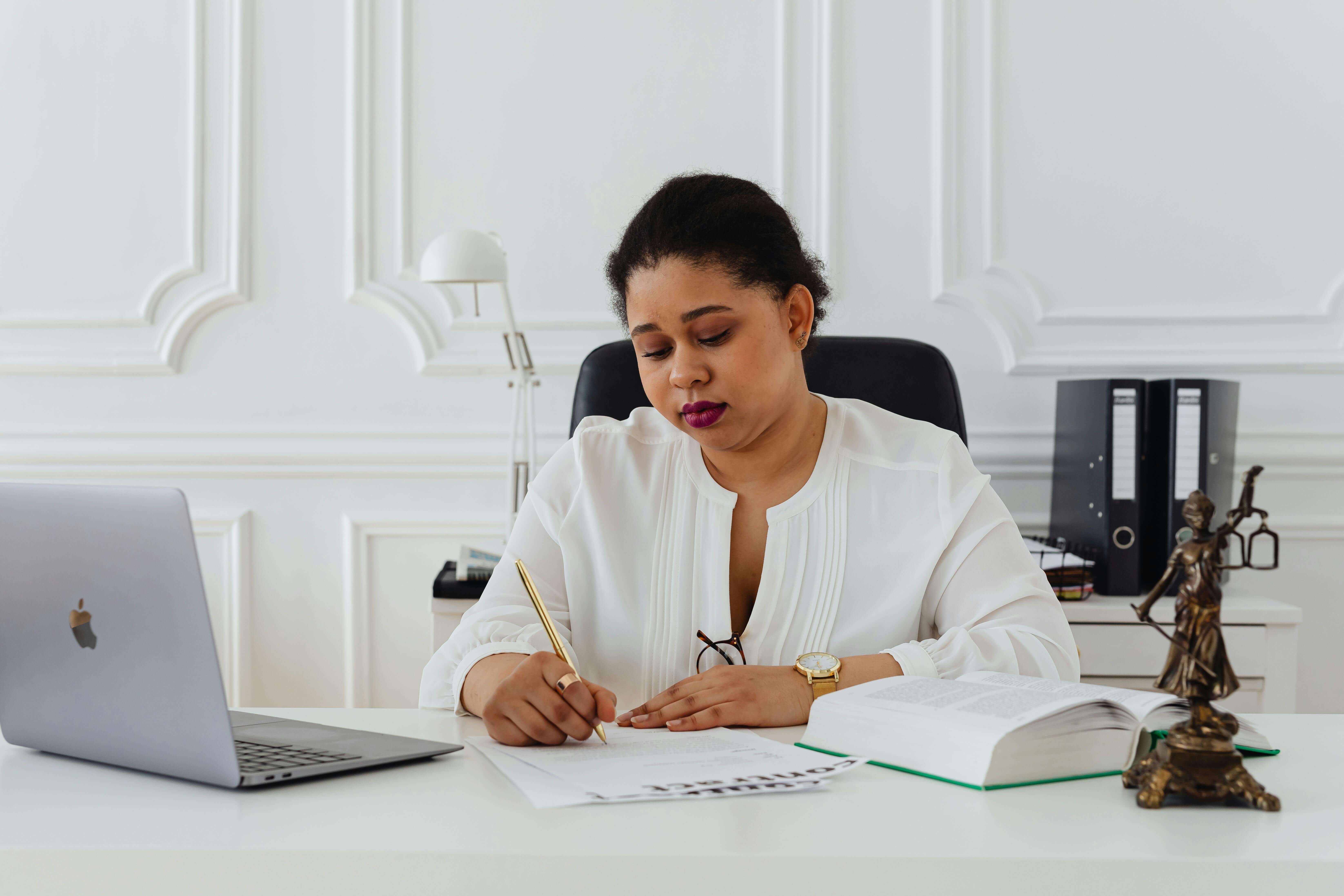 L'image montre une femme assis dans son bureau, où l'on peut voir un ordinateur et une statue en bronze de la justice, en train de signer un document  juridique.