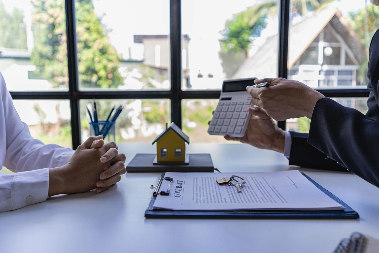 Une personne en costume utilisant une calculatrice pour expliquer la rentabilité d'un investissement locatif, avec une maquette de maison jaune et un contrat posés sur la table