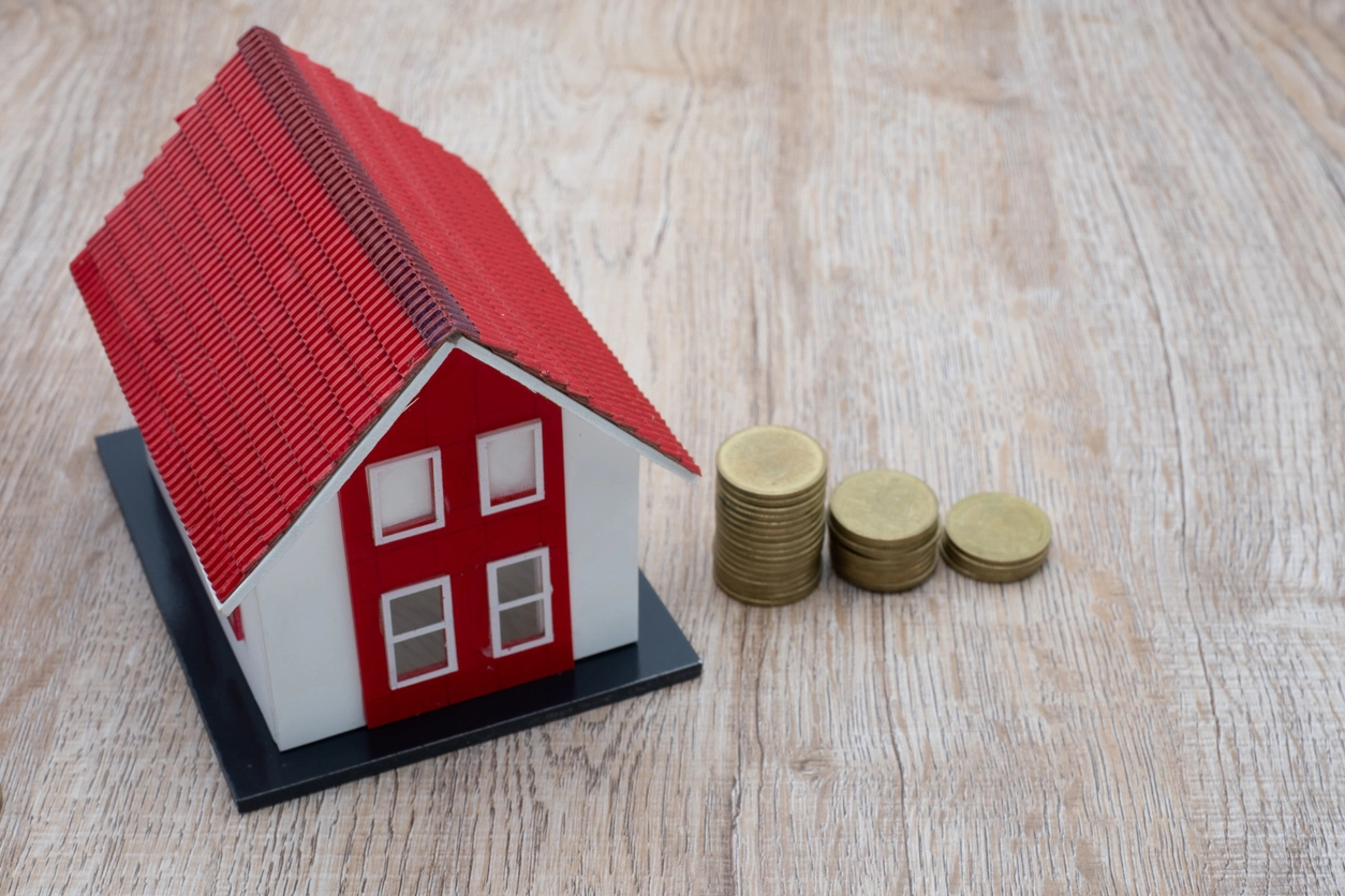 L'image représente une maison miniature blanche avec un toit rouge vif, placée sur une surface en bois. À droite de la maison, plusieurs piles de pièces de monnaie dorées sont alignées, symbolisant le coût d'une gestion locative