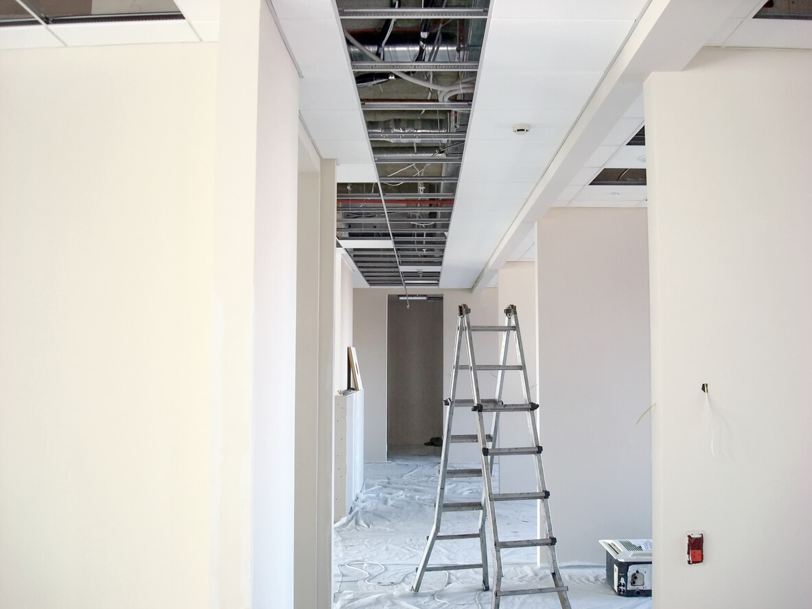 Intérieur d'un appartement en cours de rénovation. Une échelle métallique est installée au milieu de l'appartement. Au fond du couloir de l'appartement, il y a une porte grise.