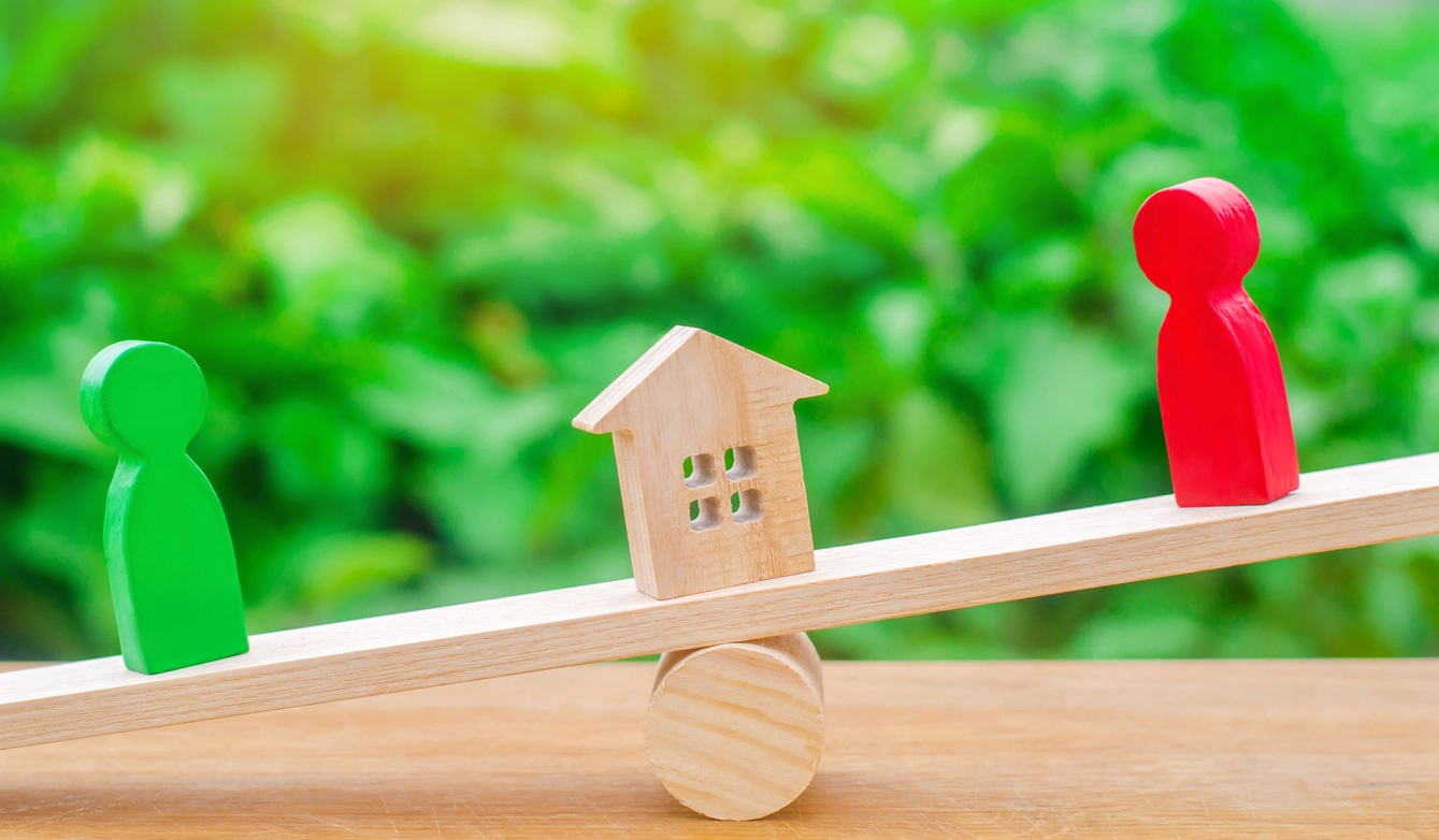 Équilibre précaire entre une maison en bois (nue-propriété) et deux personnages (usufruitiers), symbolisant le partage des droits sur un bien immobilier