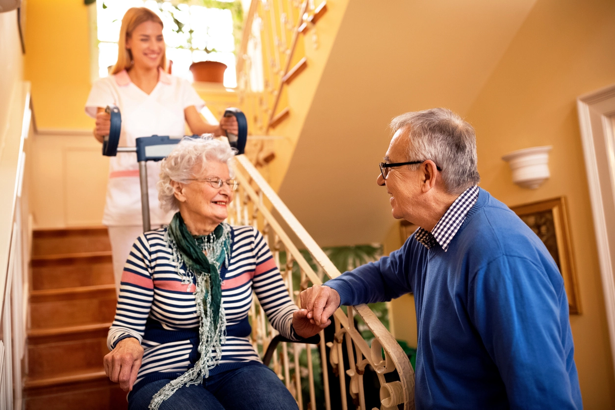 Un couple de seniors souriant chaleureusement, avec une femme assise sur un monte-escalier assistée par une aide-soignante en arrière-plan