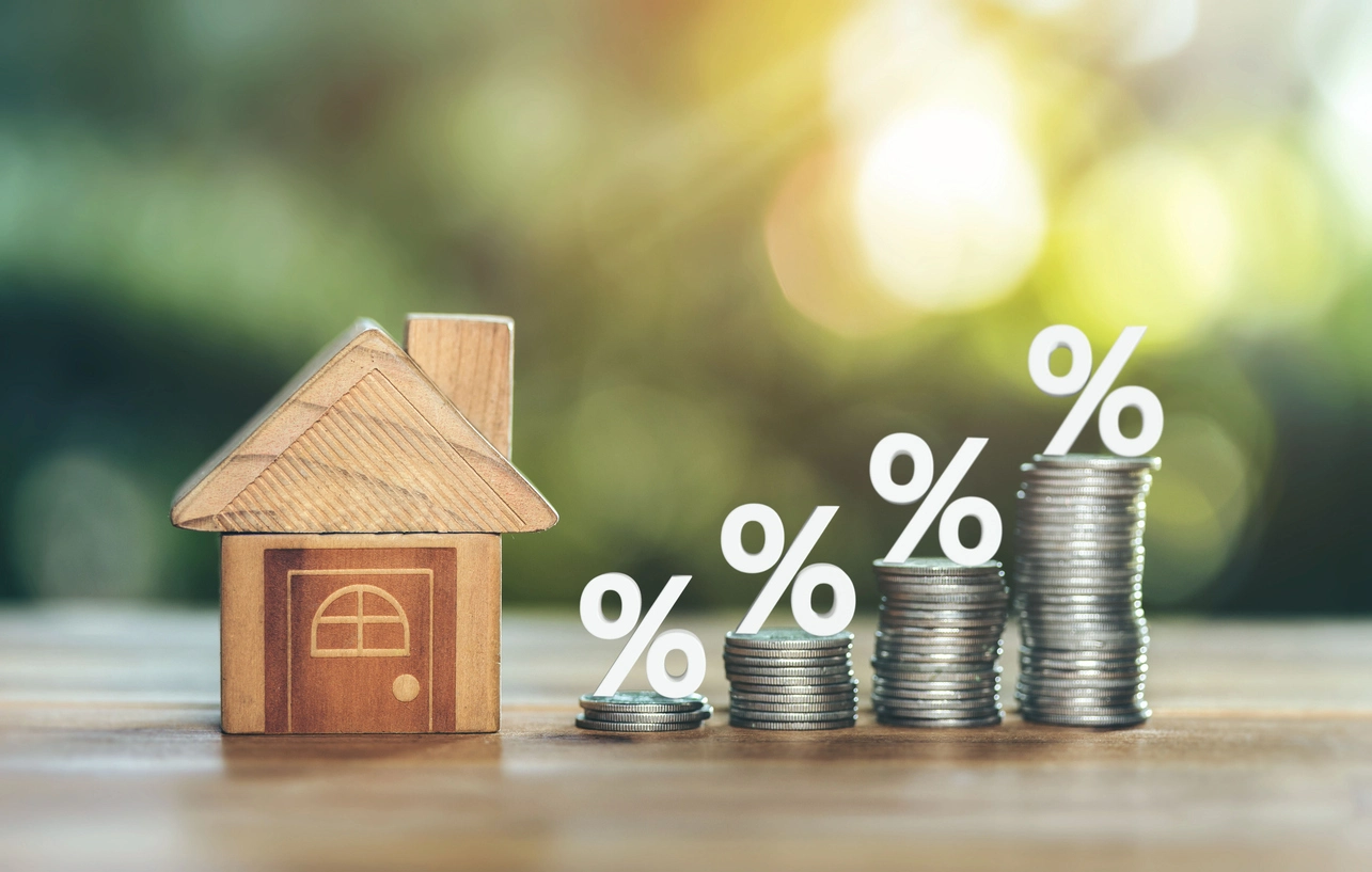 Maison en bois à côté de piles de pièces croissantes avec des symboles de pourcentage, représentant l'augmentation des taux d'intérêt dans l'immobilier.