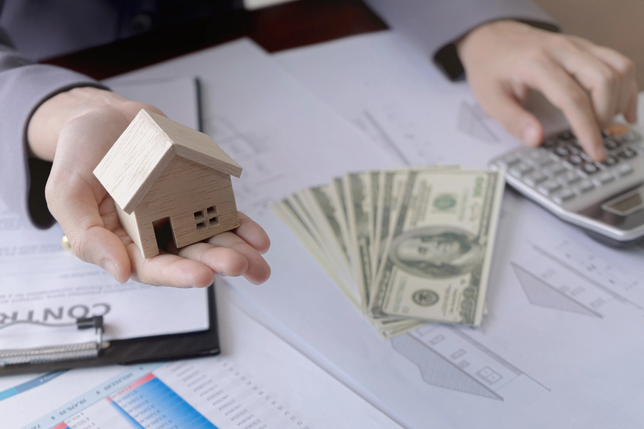 Main tenant une maison en bois miniature, avec des billets de dollars et une calculatrice sur des documents financiers, illustrant un prêt immobilier.