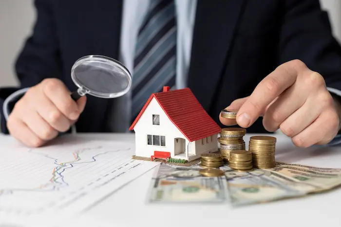 Personne en costume manipulant des pièces devant une maquette de maison et des billets, image illustrant la fiscalité immobilière et la gestion des impôts fonciers.