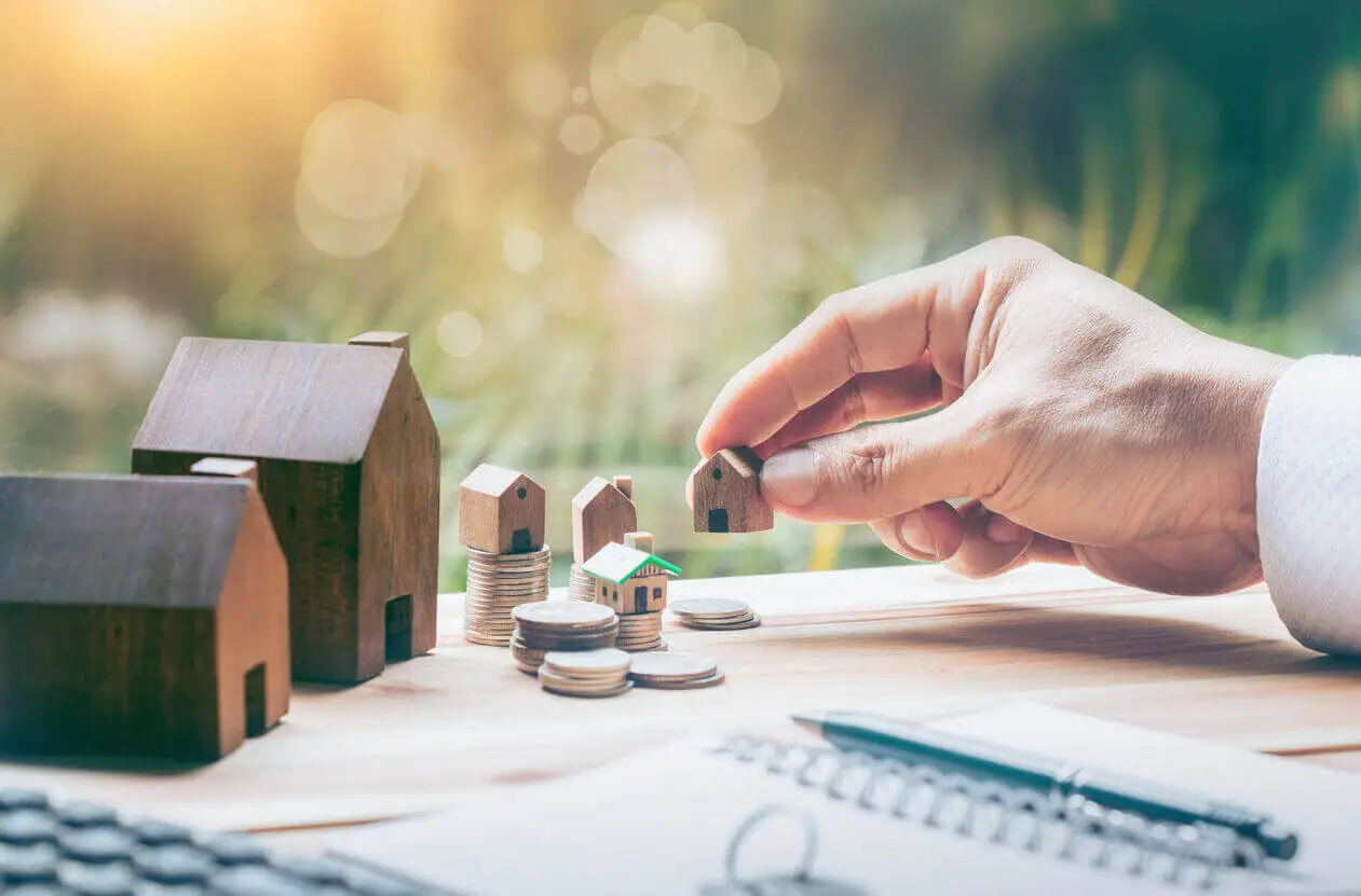 Une main tient une petite maison en bois au-dessus de piles de pièces, entourée de modèles de maisons. L’image symbolise l’investissement et la croissance immobilière.