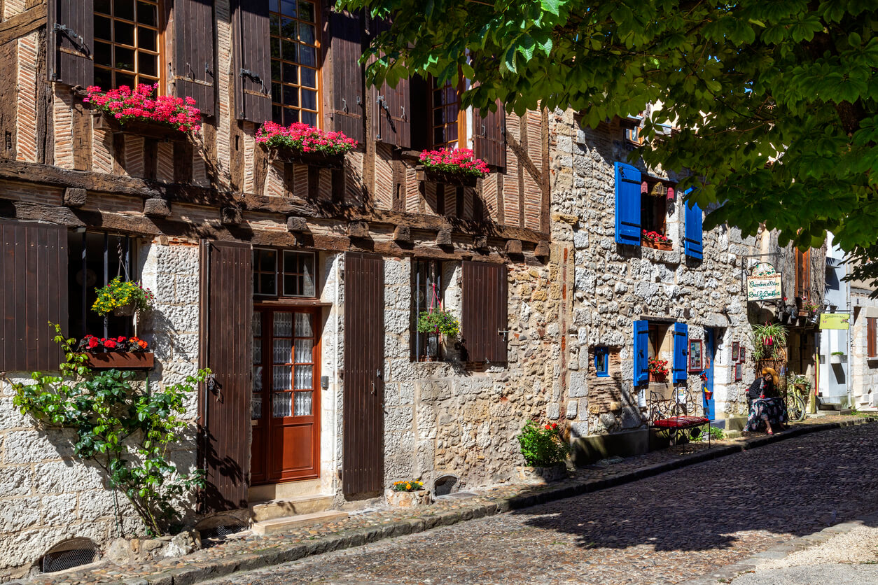 Façades de maisons anciennes dans le centre historique de Bergerac, avec des murs en pierre et des colombages en bois. Des volets en bois foncé encadrent les fenêtres ornées de jardinières fleuries. La rue pavée ajoute au charme authentique de cet ensemble architectural. À droite, on peut voir des volets bleus lumineux sur une maison en pierre.