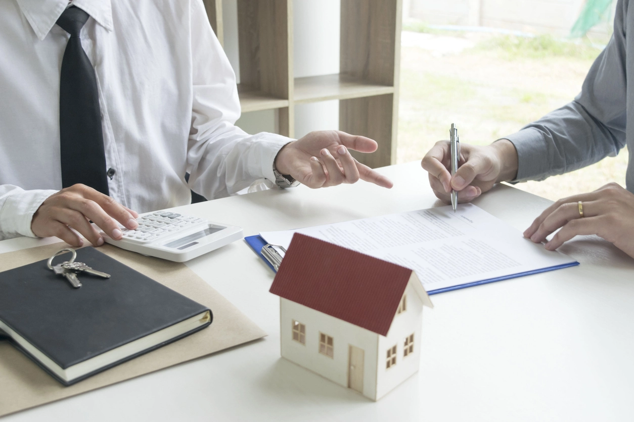 Deux personnes discutent d’un contrat, accompagné d’une maquette de maison, d’une calculatrice et de clés. Cette scène illustre la résiliation d’un contrat de gestion locative due à des fautes du mandataire de gestion locative