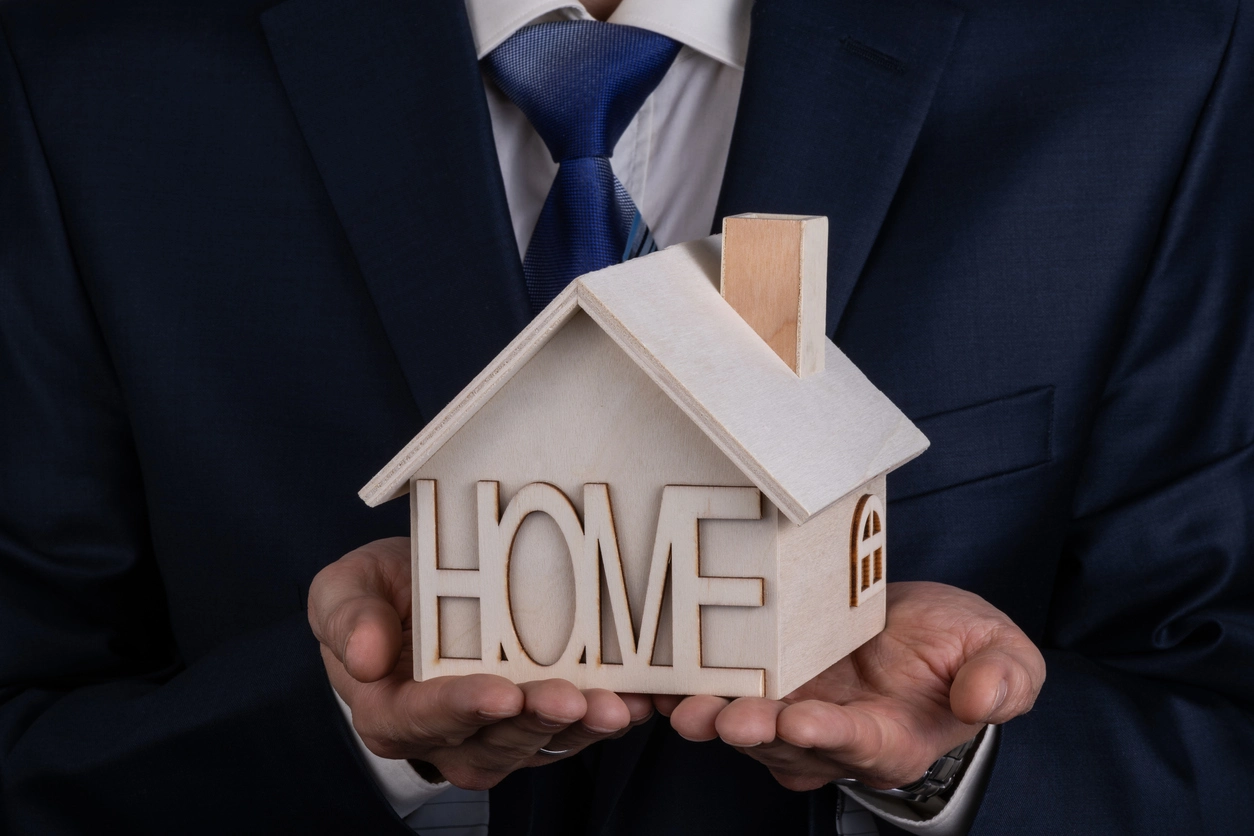 Un homme en costume tient une maison en bois avec l'inscription 