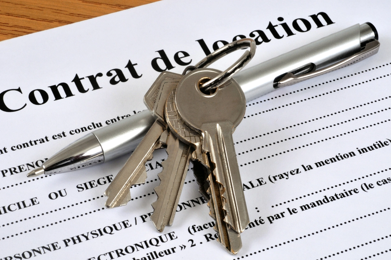 Des clés posées sur un stylo argenté, le tout placé sur un contrat de location imprimé en noir sur une feuille blanche, visible sur une table en bois clair