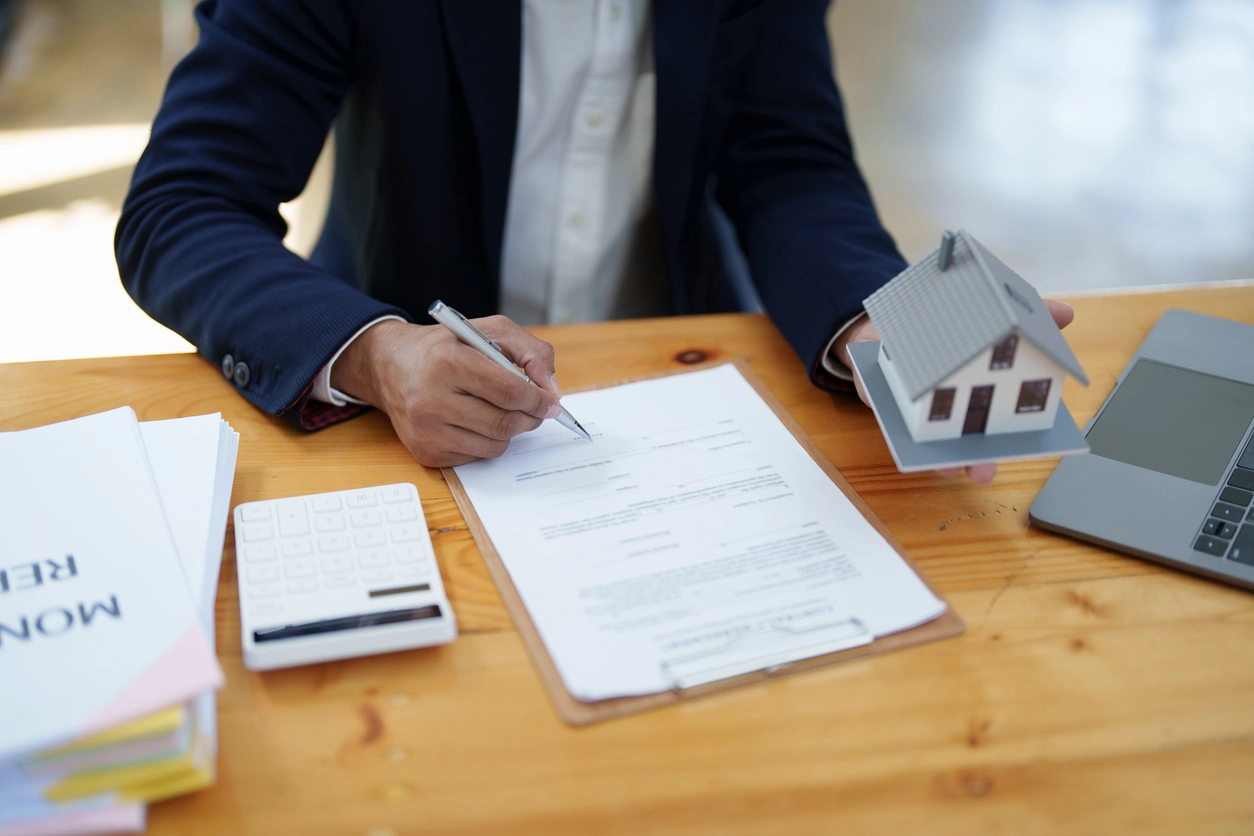 Un professionnel en costume signe un contrat sur un bureau en bois tout en tenant une maquette de maison, symbolisant un accord immobilier ou une gestion locative Tours.