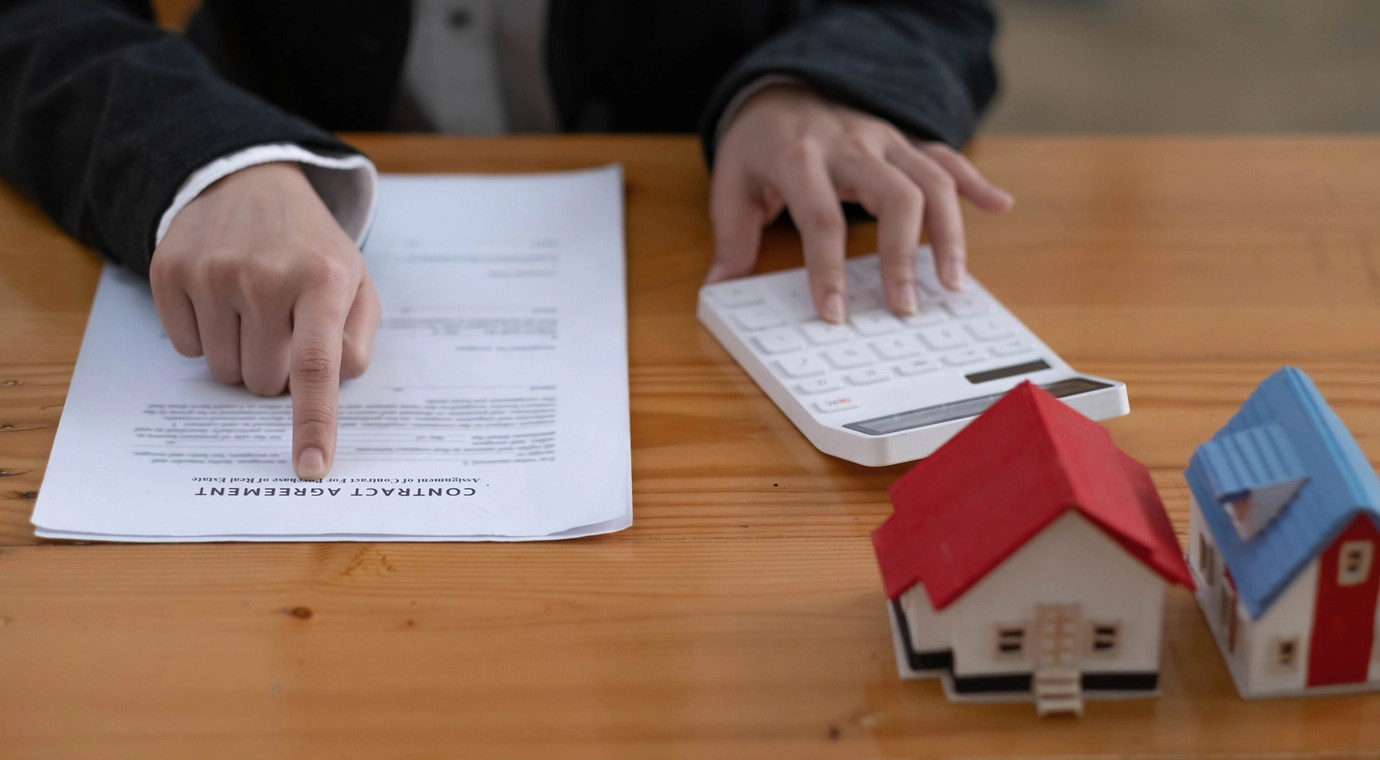 Une personne montre un contrat de bail sur une table en bois, avec une main sur une calculatrice et deux petites maquettes de maisons en arrière-plan