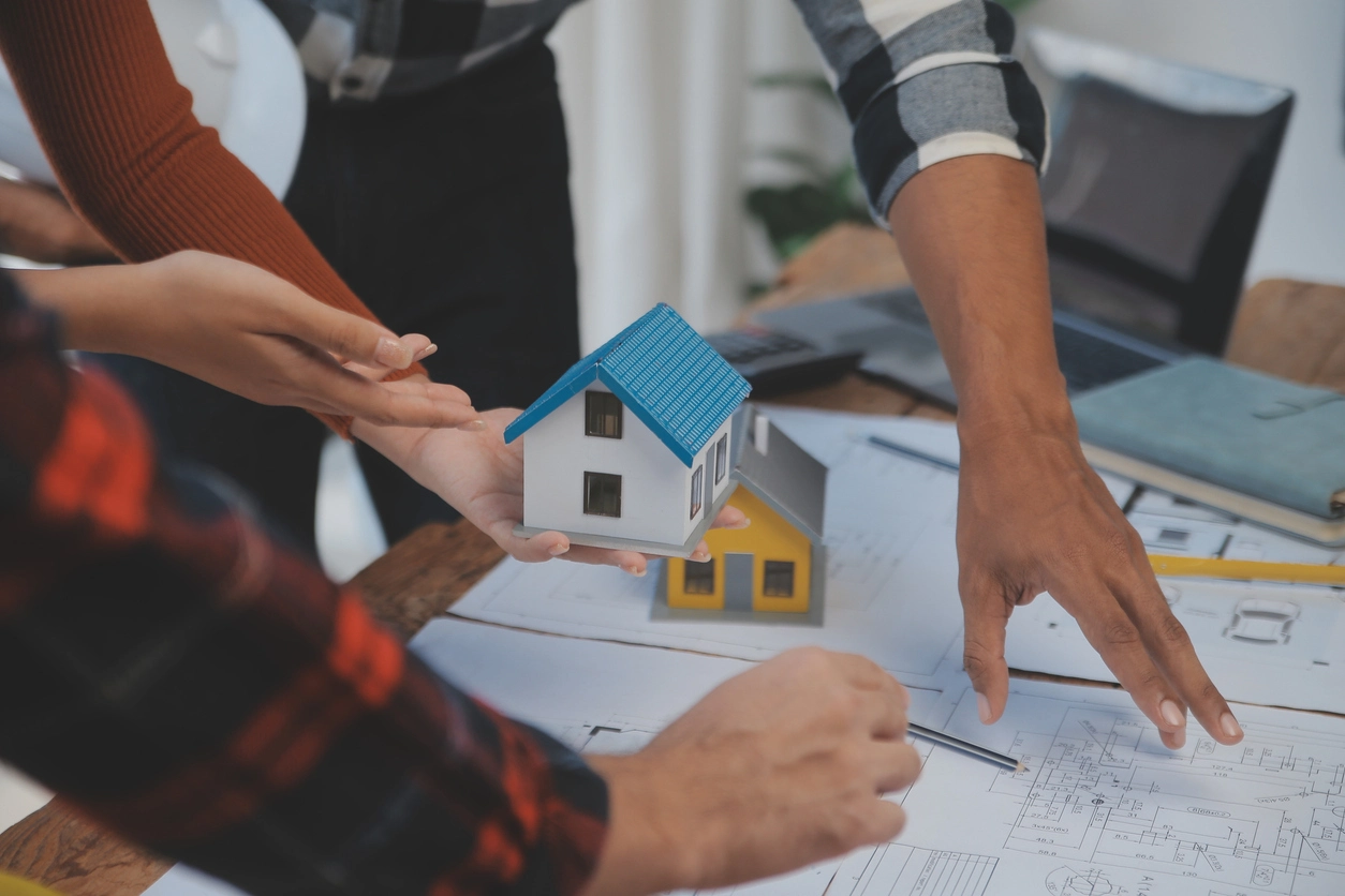 Des personnes analysent des plans architecturaux sur une table. Une main tient une maquette de maison avec un toit bleu. L’image illustre l’étude un projet immobilier avec 30 000 euros.