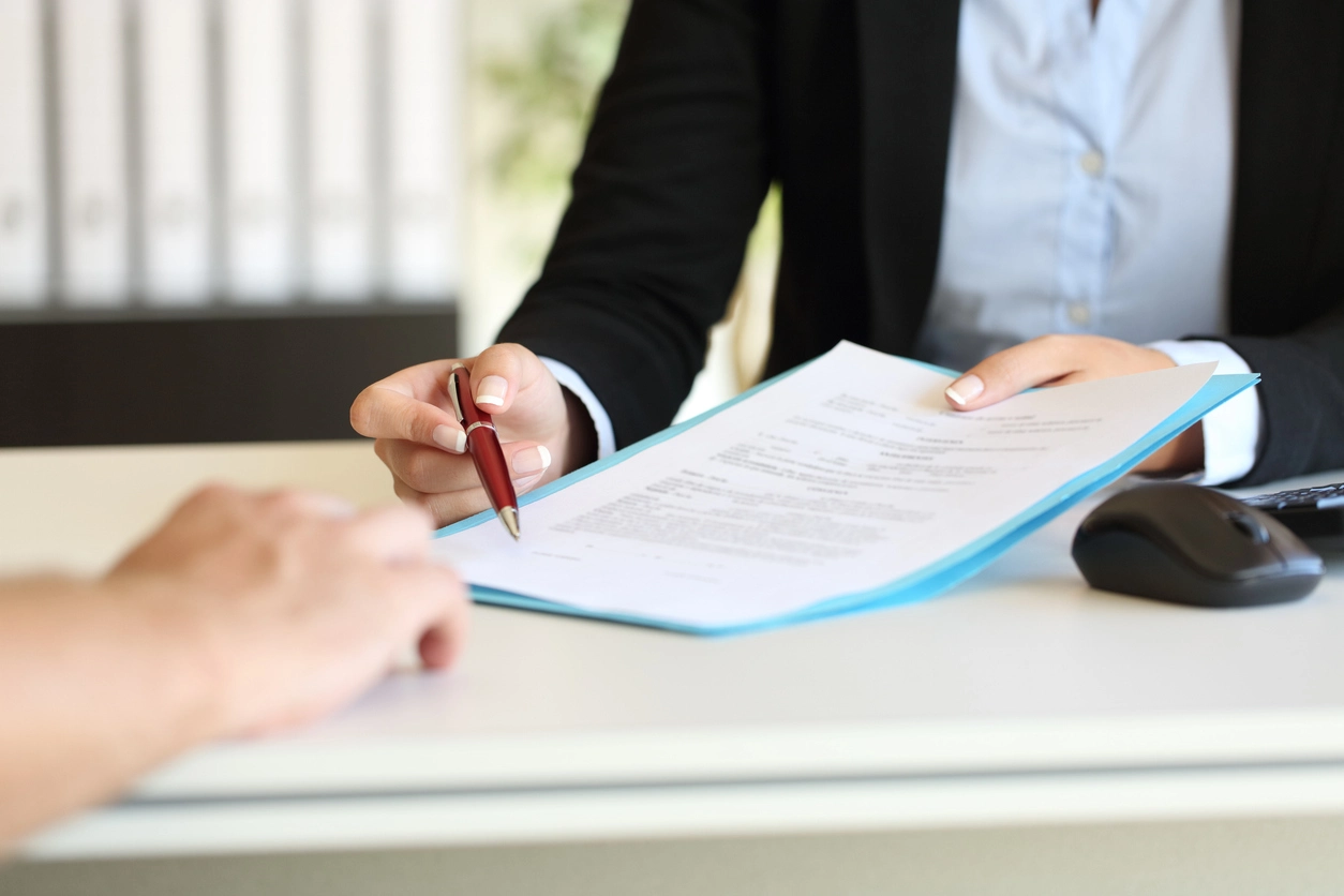 Une personne en costume tenant un stylo rouge et présentant un contrat de gestion locative. Le document est placé dans une chemise bleue, tandis qu'une autre personne tend la main pour interagir