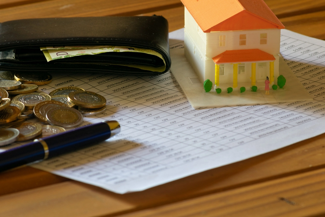 Une table en bois avec une maquette de maison, un portefeuille ouvert contenant des billets, des pièces de monnaie dispersées, un stylo et une feuille imprimée. L'image illustre la réalisation d'un devis gestion locative