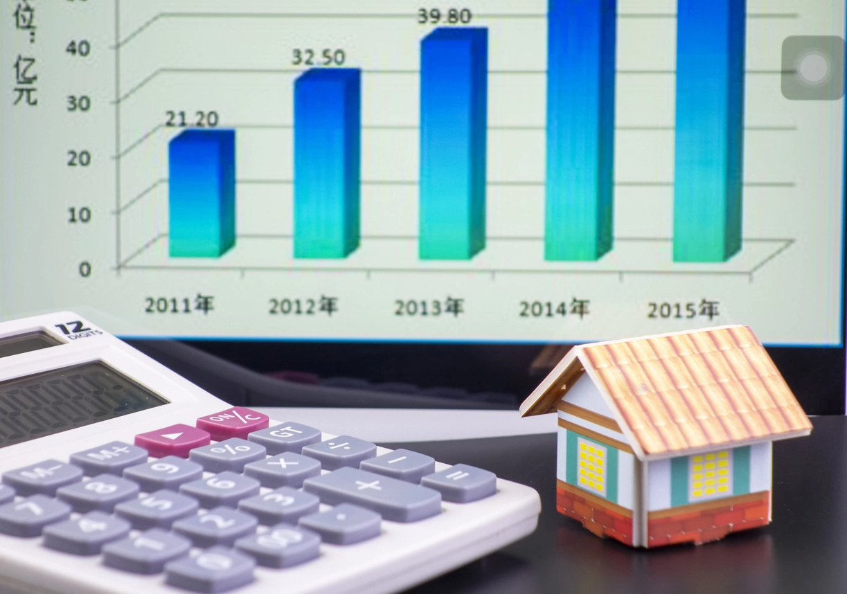 Sur un bureau, une calculatrice blanche affiche des chiffres, à côté d'une petite maison en carton colorée. Un écran d'ordinateur en arrière-plan montre un graphique à barres en augmentation illustrant la rentabilité nette d'un investissement locatif