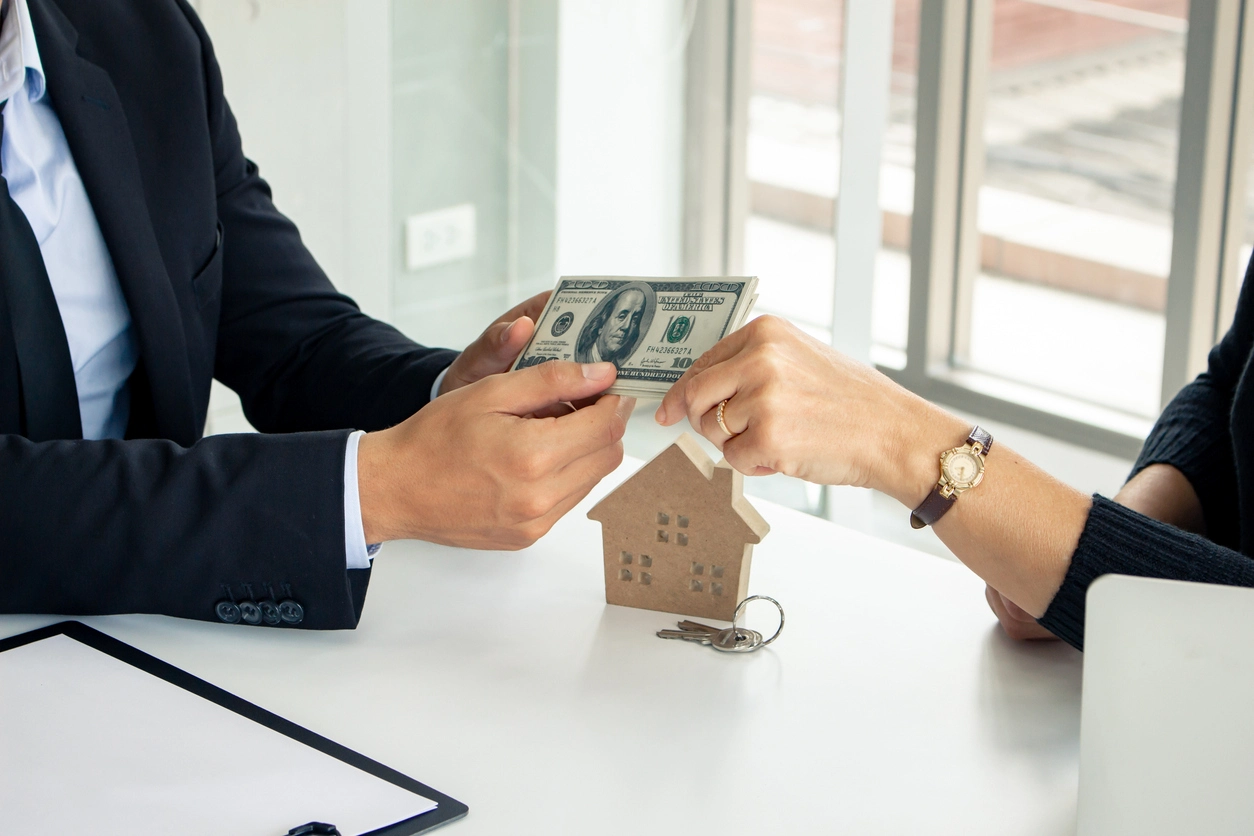 Une transaction financière entre deux personnes, avec un échange de billets de banque devant une maquette de maison en bois et un trousseau de clés, symbolisant un tarif de gestion locative Tours