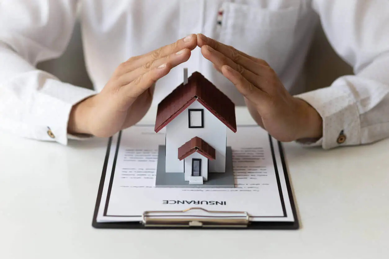 Des mains protègent une maquette de maison posée sur un contrat d’assurance. L’image symbolise la protection et la sécurité offertes par une assurance habitation.