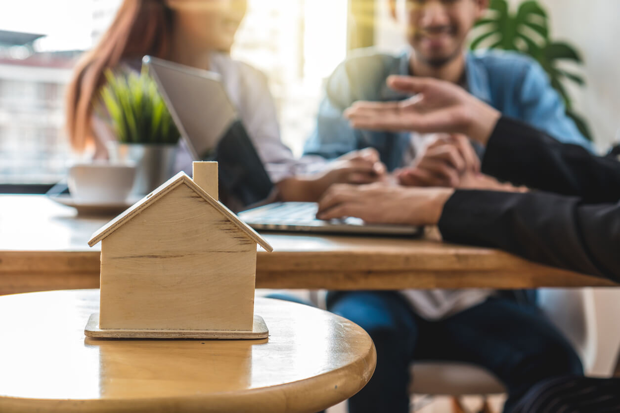 Illustration représentant une petite maison au premier plan, symbole de patrimoine, tandis qu'en arrière-plan, des personnes discutent, évoquant la fiscalité et les stratégies liées à l'assurance-vie en 2024.