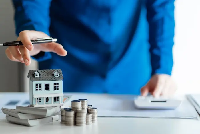 Personne en chemise bleue utilisant calculatrice avec maison miniature et pièces empilées, image illustrant le simulateur de rentabilité d’investissement immobilier.