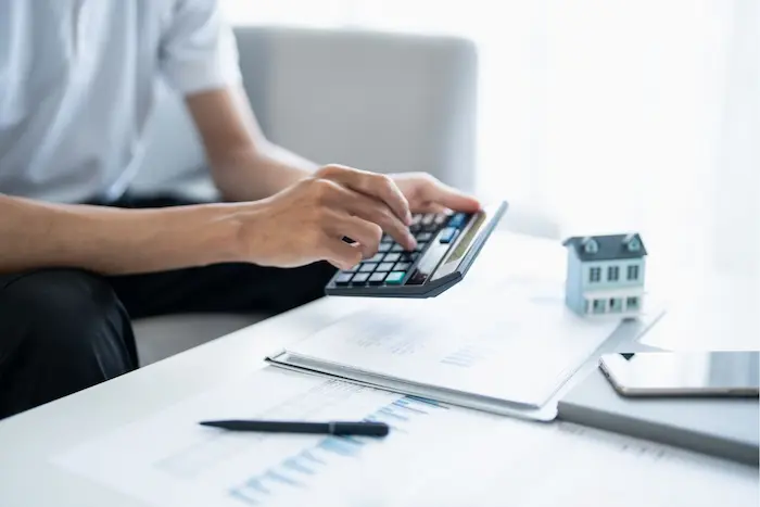 Personne utilisant une calculatrice avec documents financiers et maquette de maison, image qui illustre le calcul du rendement locatif.