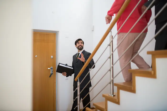 Agent en costume présentant un escalier moderne à des visiteurs, scène typique d’une visite de bien immobilier.