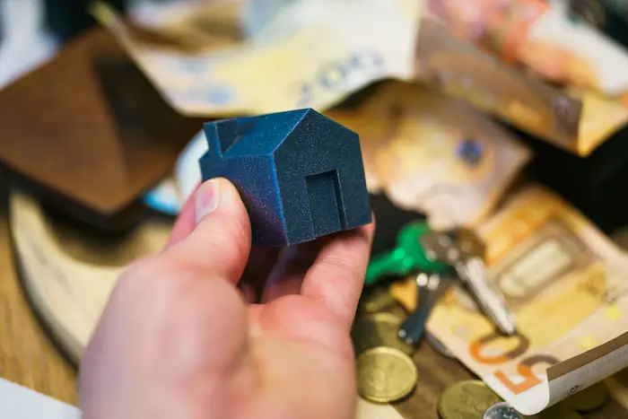 Main tenant une maison miniature bleue avec billets et pièces en arrière-plan, reflet du taux d’endettement lié au crédit immobilier.