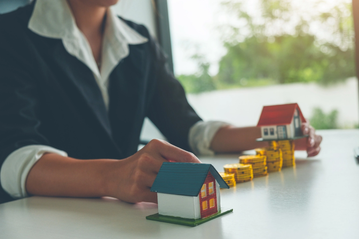 Une personne en costume manipule des miniatures de maisons sur un bureau, accompagnées de piles de pièces dorées, symbolisant la gestion locative Angers et l'investissement immobilier