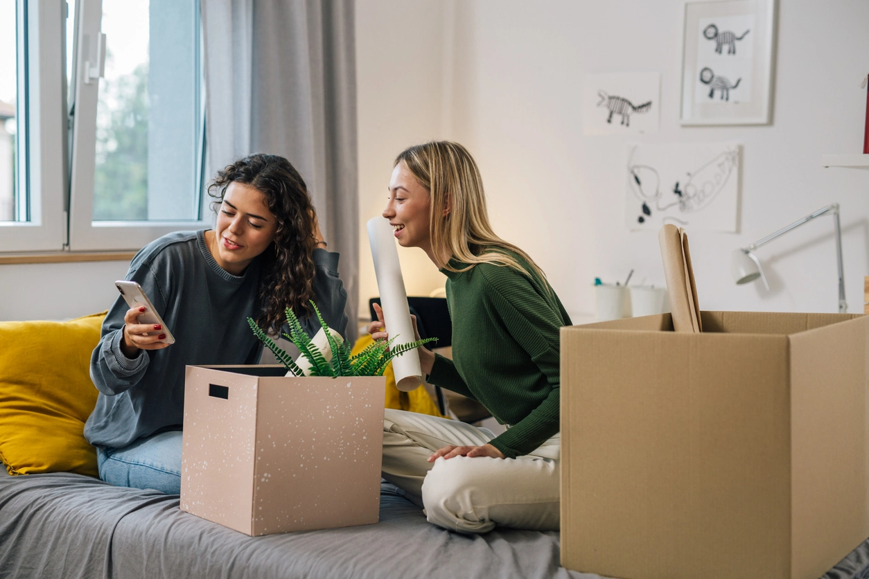 Deux étudiantes installées dans une chambre décorée, discutant avec enthousiasme. Elles organisent leur espace de vie dans une résidence étudiante accueillante et lumineuse