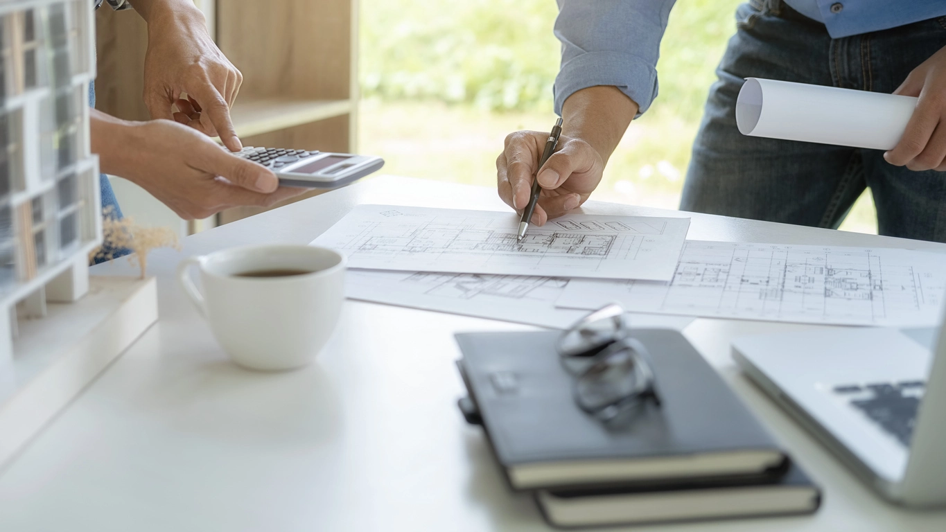 Deux professionnels analysent des plans architecturaux sur une table, avec une calculatrice, un carnet, une tasse de café et une maquette de bâtiment à proximité. L’image illustre un projet dans une SCPI