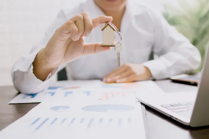 Personne tenant une clé en forme de maison devant graphiques financiers, reflet d’une simulation de prêt achat-revente immobilier.