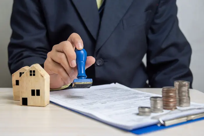 Personne en costume tamponnant un document officiel avec maisons miniatures et pièces empilées, image illustrant la validation d’un dossier de prêt immobilier.
