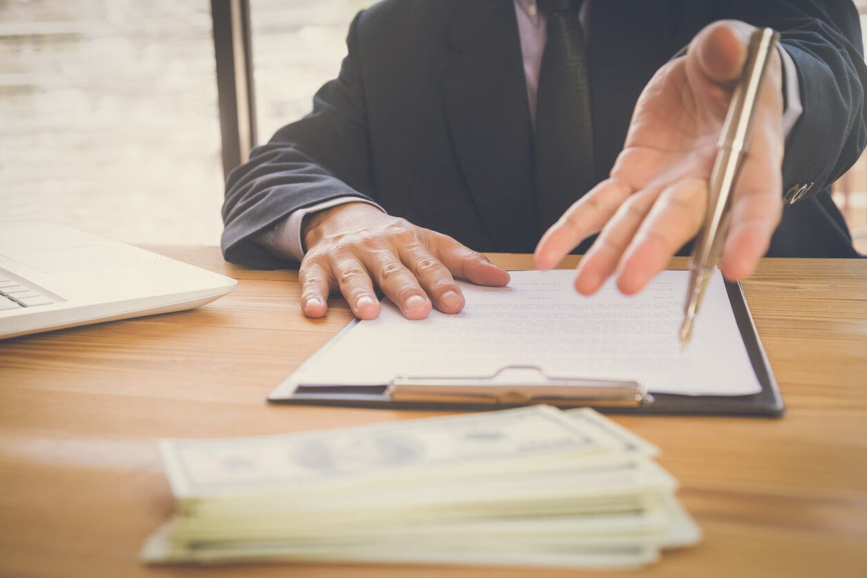 une main tendant un stylo pour signer un modèle statuts SCI. Sur la table, il y a un ordinateur portable et des billets de banque