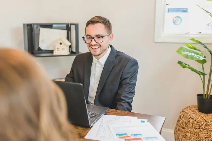 Un courtier prêt immobilier souriant en costume conseille une cliente dans son bureau devant son ordinateur pour obtenir le meilleur taux de financement local