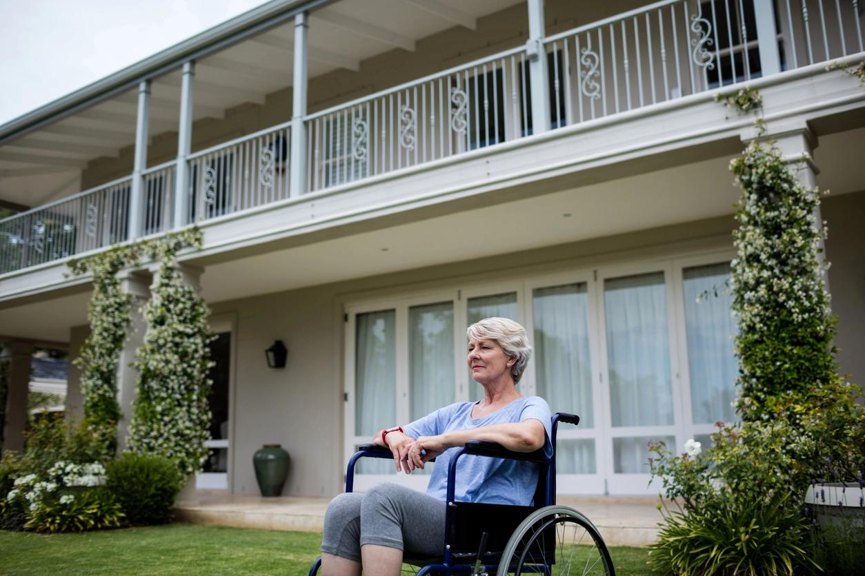 Une femme senior en fauteuil roulant assise dans un jardin bien entretenu devant une élégante résidence équipée de balcons, illustrant un cadre paisible idéal pour un investissement résidence senior