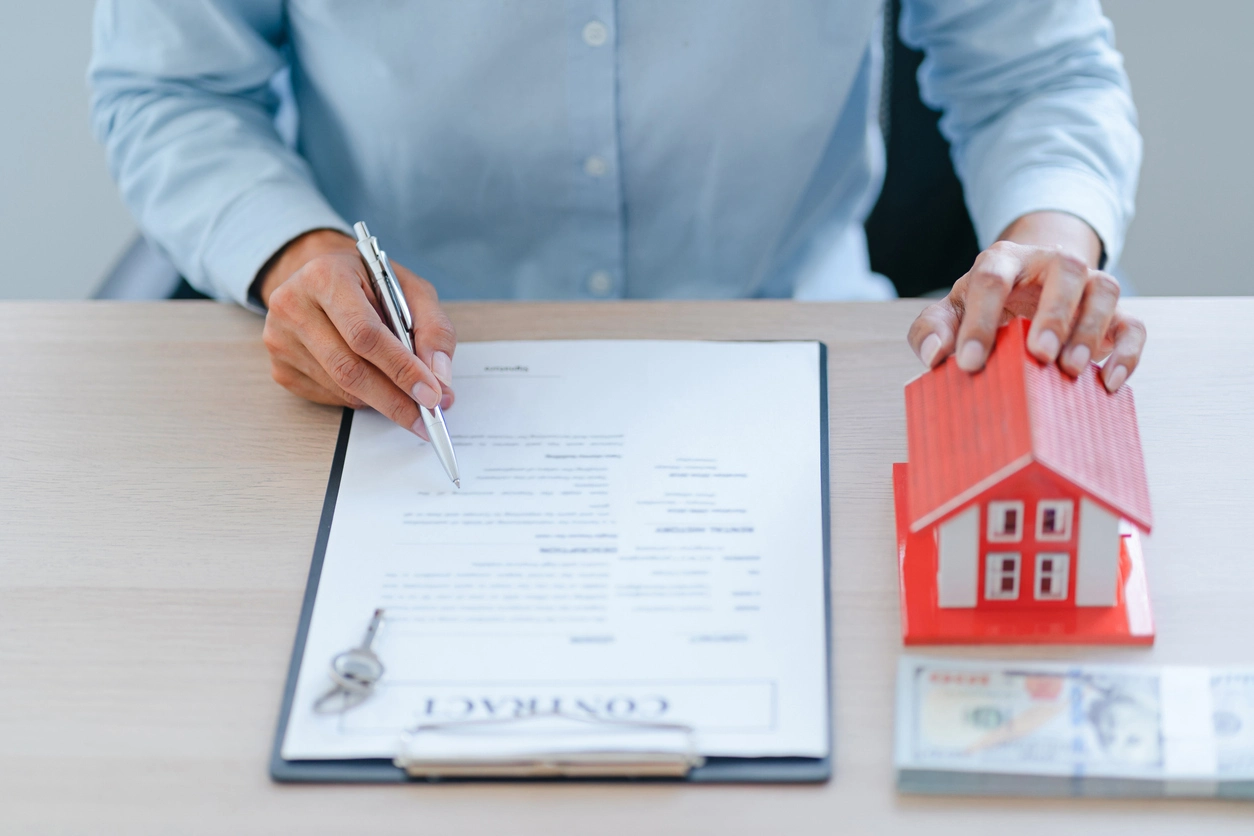 Un agent immobilier signe un contrat, tenant un stylo, avec une miniature de maison rouge, des clés et des billets de banque posés sur la table