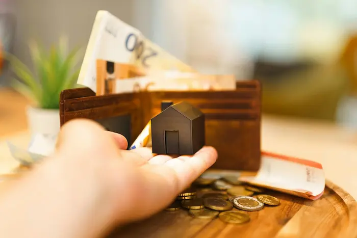 Main tenant une maison miniature noire devant portefeuille rempli de billets et pièces en euros illustrant le principe du prêt relais immobilier.